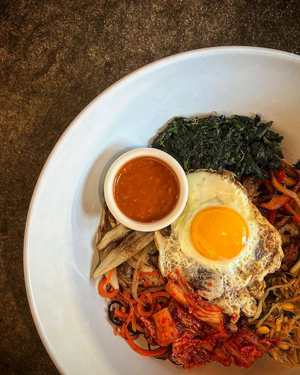 A dish featuring a sunny-side-up egg, sautéed vegetables, greens, kimchi, and a small container of sauce is presented in a white bowl.