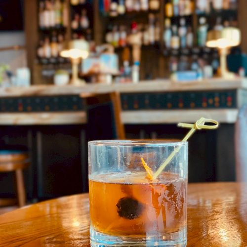 A cocktail garnished with a cherry and an orange peel on a wooden table with a blurred bar background, featuring shelves of bottles and warm lighting.