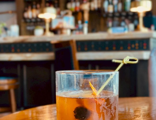 A cocktail garnished with a cherry and an orange peel on a wooden table with a blurred bar background, featuring shelves of bottles and warm lighting.