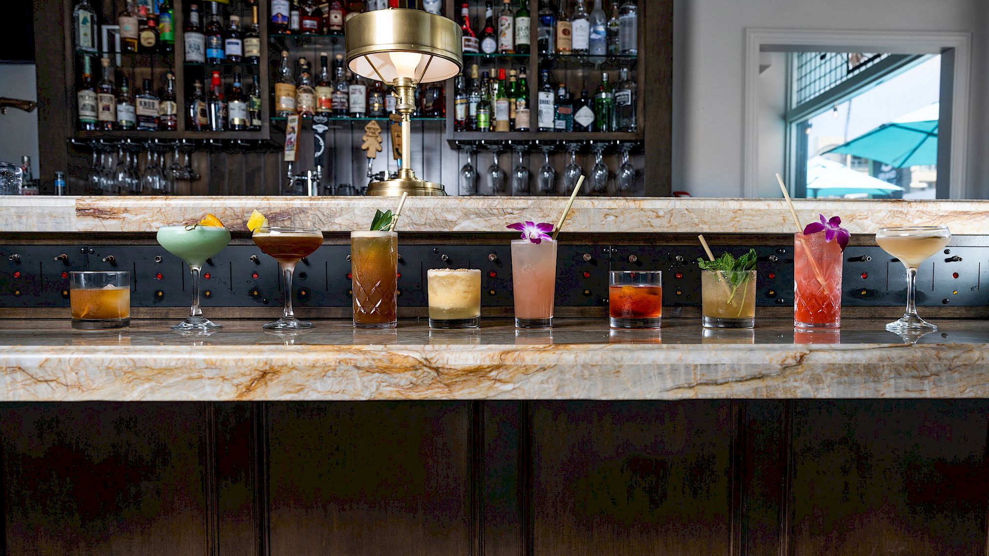 The image shows a row of diverse cocktails on a marble bar counter, with a well-stocked bar and lamp in the background.