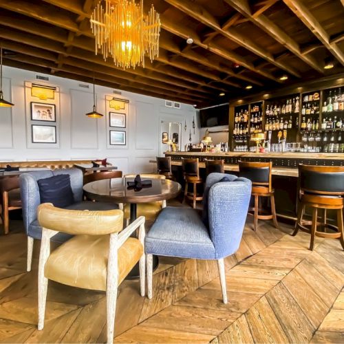 A cozy, modern bar with wooden decor, a well-stocked bar area, a mix of chairs and tables, pendant lighting, and a stylish chandelier ending the sentence.