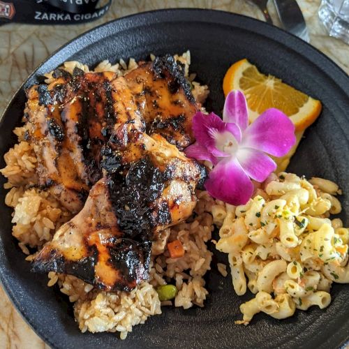 A plate with grilled chicken on rice, a serving of macaroni salad, a slice of orange, and an edible flower for garnish.