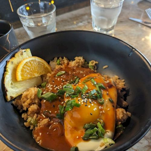 The image shows a bowl of food containing eggs, greens, a slice of lemon, and sauce, along with two glasses of water on a table.