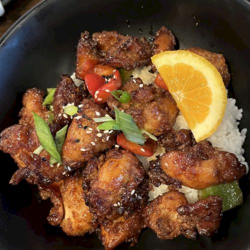 A dish with rice, fried chicken pieces, garnished with green onions, sesame seeds, red peppers, and an orange slice, served in a black bowl.