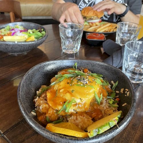 The image shows a meal on a wooden table, featuring a bowl of food topped with sauce and herbs, a salad, glasses of water, and a person eating.