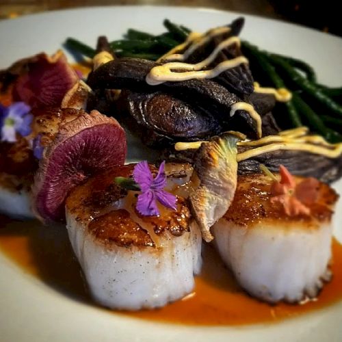 The image shows a gourmet plate with seared scallops, garnished with colorful edible flowers, vegetables, and a sauce, arranged artfully.