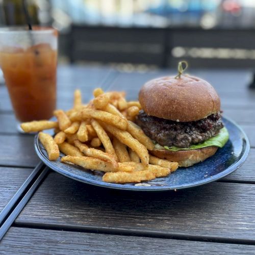 A plate with a burger and fries on a wooden table, accompanied by a drink in a glass with ice and a straw, beside another glass and sunglasses.