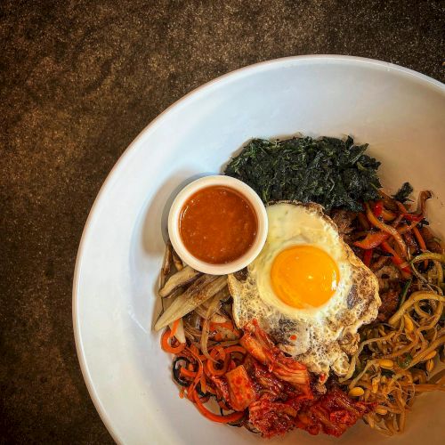 A dish with a fried egg on top, assorted vegetables, greens, and a small bowl of sauce on the side, served in a white bowl.