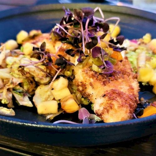 The image shows a plate of breaded fish garnished with microgreens and served alongside mixed diced vegetables and shredded salad.