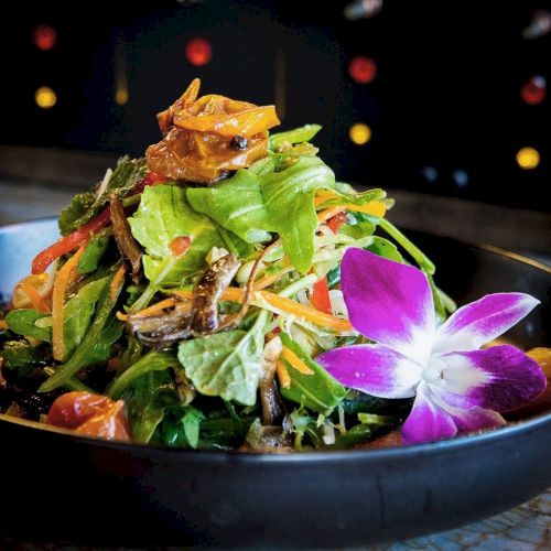 A vibrant salad with mixed greens, shredded vegetables, topped with an edible flower, served in a black bowl, garnished with a colorful purple flower.