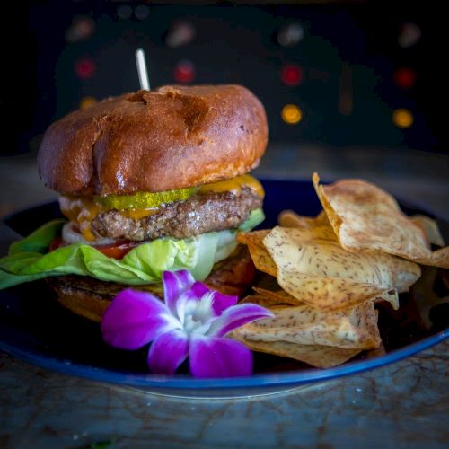 A burger with lettuce, tomato, and cheese, served with crispy chips and garnished with a purple orchid flower on a dark plate.