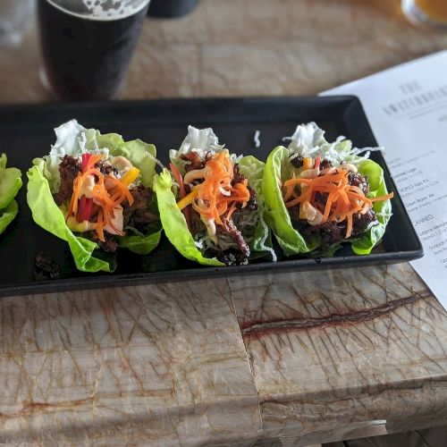 The image shows a rectangular black plate with four lettuce wraps filled with vegetables and meat, placed on a marble surface next to a drink and menu.