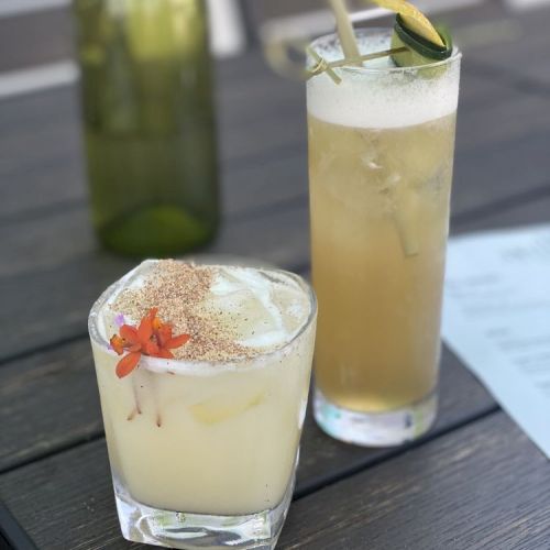 Two mixed drinks on a dark wooden table; one short glass garnished with a flower, the other tall glass garnished with a lime wedge and a straw.