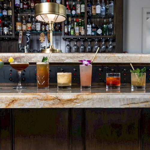 A variety of colorful cocktails are displayed on a marble bar counter in front of a well-stocked back bar with a lamp in the center.