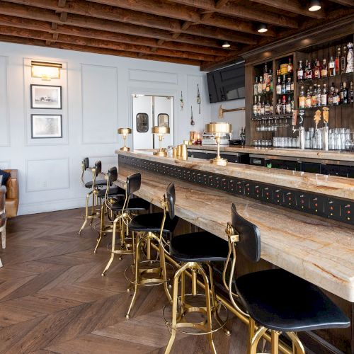 The image shows a stylish bar with a wooden ceiling, a long counter with bar stools, a seating area with a couch and chairs, and a well-stocked bar.