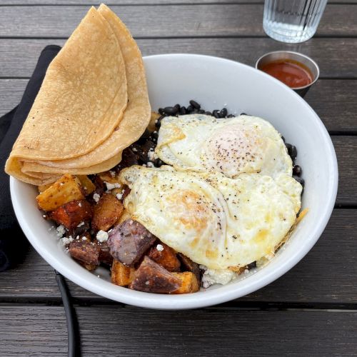 A bowl containing rice, roasted vegetables, and two fried eggs, accompanied by corn tortillas and a glass of water on a wooden table.