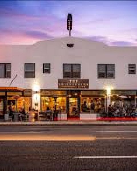 The image shows a street view of a well-lit, white building housing a restaurant and cafe, with cars passing by in front at dusk.