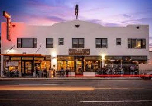 The image shows a street view of a well-lit, white building housing a restaurant and cafe, with cars passing by in front at dusk.