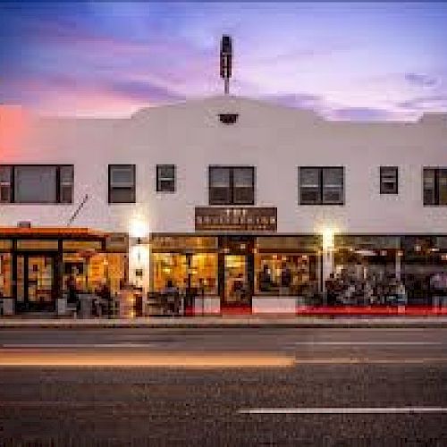 The image shows a street view of a well-lit, white building housing a restaurant and cafe, with cars passing by in front at dusk.